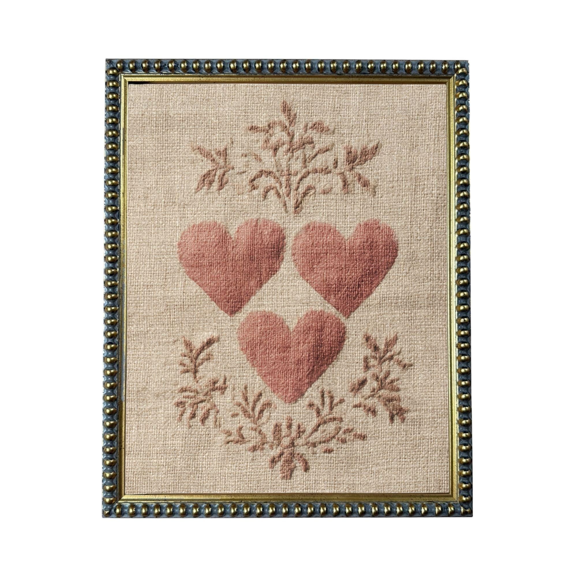 a framed piece of artwork with a beige background and a border. The artwork is a close-up of three red hearts arranged in a triangular formation, surrounded by a floral design.